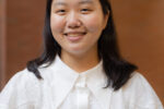 Headshot of a person smiling, wearing a white shirt with an embroidered pattern, against a blurred brown background.