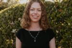 A person with curly hair is smiling and standing in front of a sunlit green hedge. The individual is wearing a black top and a necklace.