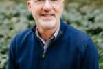 Headshot of a person with short white hair and glasses, wearing a navy blue sweater over a checkered shirt, standing outdoors with greenery in the background.