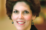 Headshot of a person with short, styled hair, wearing earrings and a dark jacket, smiling at the camera.