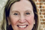 Headshot of a smiling person with long hair, wearing earrings, in front of a brick wall.
