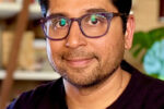 A headshot of a person with glasses, short dark hair, and facial hair, wearing a dark shirt, smiling slightly. The background features blurred shelves and a plant.