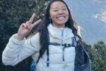 A person in a white puffy jacket with a backpack, smiling and making a peace sign with their hand. They are outdoors with a mountainous landscape in the background.