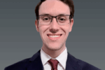 Headshot of a person wearing glasses, a dark suit, white shirt, and red tie, smiling against a plain background.