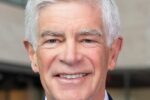 Headshot of a person with white hair, wearing a suit and tie, smiling. The background appears to be indoors.