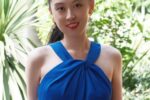 A person in a blue halter dress, wearing star-shaped earrings, stands outdoors with greenery in the background, smiling.