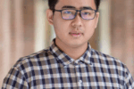 A headshot of a person wearing glasses and a plaid button-up shirt, standing in a corridor with blurred brick walls in the background.
