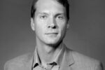 Black and white headshot of a person in a suit, looking directly at the camera with a neutral expression.