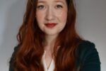 Headshot of a person with long, wavy red hair, wearing a green jacket over a light-colored top, against a neutral background.