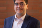 Headshot of a person wearing glasses, a dark blazer, and a white shirt, standing against a brick wall background.