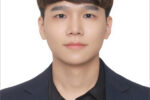 Headshot of a person with short brown hair, wearing a navy blazer and black shirt, against a plain white background.