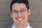 Headshot of a smiling person with short dark hair, wearing glasses and a white shirt, in front of a brick wall.