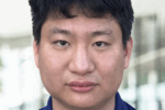 A headshot of a person with short, dark hair wearing a blue collared shirt, set against a blurred background.