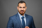 A headshot of a person wearing a blue suit, light blue shirt, and blue tie against a gray background.